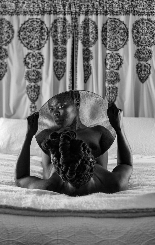Zanele Muholi liegt in diesem Selbstporträt auf einem Bett und betrachtet mit durchdringendem Blick das eigene Spiegelbild mit einem runden Spiegel in den Händen. Die Glanzlichter in dieser Fotografie sind bewusst abgeschwächt, was die dunkle Haut Muholi’s markanter wirken lässt.